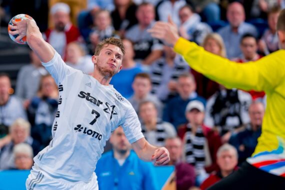 Ein Handballspieler in einem weißen Trikot bereitet sich auf einen Schuss vor, während er einem Torwart in einem gelben Trikot gegenübersteht, während im Hintergrund eine Menge von Zuschauern zusieht.