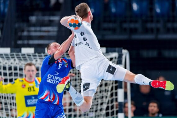 Ein Handballspieler in einem weißen Trikot springt zum Ball, während ein Verteidiger in einem blauen Trikot versucht, ihn zu blockieren. Ein Torwart in Gelb steht vor dem Tor bereit.