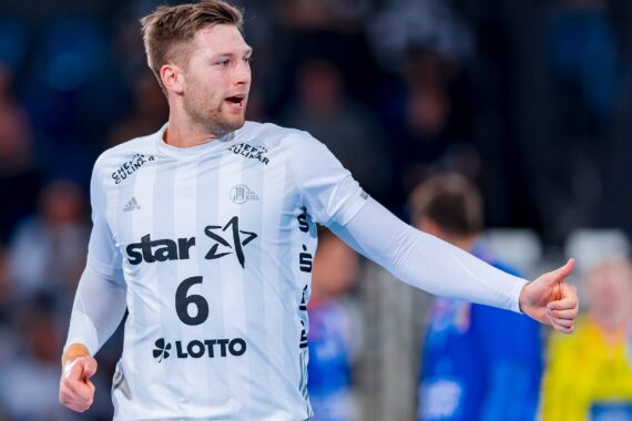 Ein männlicher Handballspieler in einem weißen Trikot mit der Nummer 6 und dem Stern- und LOTTO-Logo gestikuliert mit seiner rechten Hand während eines Spiels. Der Hintergrund zeigt unscharfe Spieler und Zuschauer.