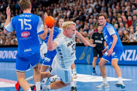 Ein Handballspieler in einem weißen Trikot springt mit einem orangefarbenen Ball nach vorne und bereitet sich auf einen Schuss vor, während ein Verteidiger in einem blauen Trikot versucht, ihn abzuwehren. Das Spiel findet in einer Halle mit Zuschauern im Hintergrund statt.