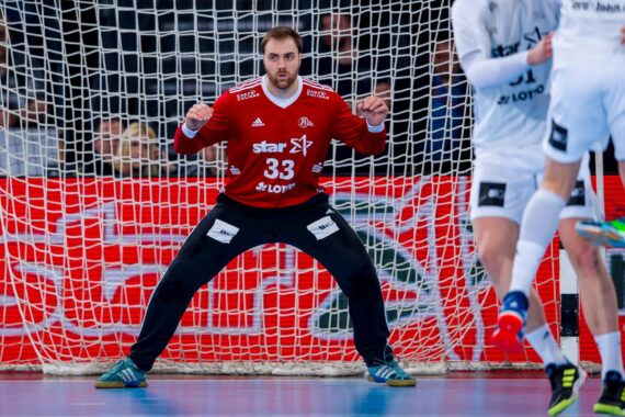 Ein Handballtorwart in einem roten Trikot mit der Nummer 33 steht wachsam vor dem Tor und bereitet sich darauf vor, einen Schuss abzublocken, während sich gegnerische Spieler in weißen Trikots nähern.