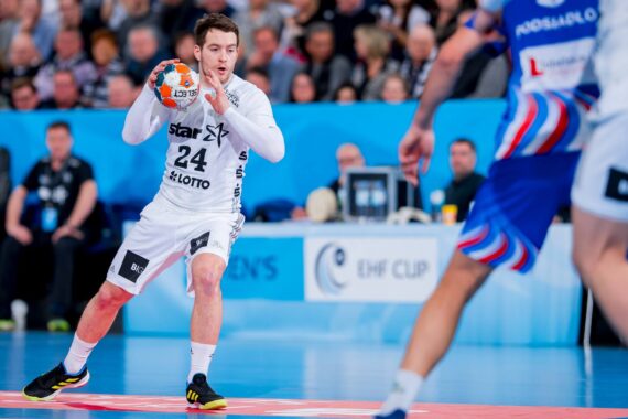 Ein männlicher Handballspieler in einem weißen Trikot, der einen orange-blauen Ball in der Hand hält, bereitet sich auf einen Pass oder einen Schuss während eines Spiels vor. Ein Verteidiger in Blau steht in der Nähe, während im Hintergrund eine Menschenmenge zusieht.