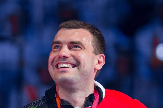 Ein Mann in einer rot-schwarzen Jacke lächelt strahlend und blickt nach oben. Um seinen Hals trägt er eine Medaille. Der Hintergrund ist unscharf mit blauen und weißen Tönen.