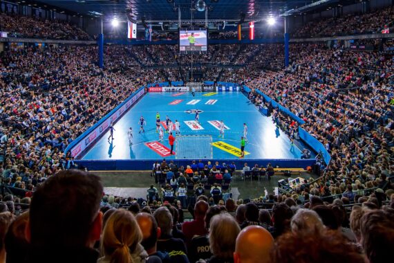 Eine große Halle, gefüllt mit Zuschauern, die ein Handballspiel auf einem hellblauen Spielfeld verfolgen, mit Mannschaften in roten und blauen Trikots und Schiedsrichtern in Neongrün.