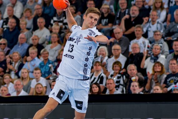 Ein männlicher Handballspieler in einem weißen Trikot springt, um einen orangefarbenen Ball zu werfen, während im Hintergrund ein Publikum von der Tribüne aus zuschaut.