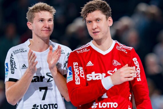 Zwei männliche Handballspieler in Mannschaftsuniformen, einer in Weiß, der klatscht, der andere in Rot, gehen nach vorne. Beide haben kurzes, hellbraunes Haar. Der Hintergrund ist unscharf und deutet auf eine Sporthalle hin.