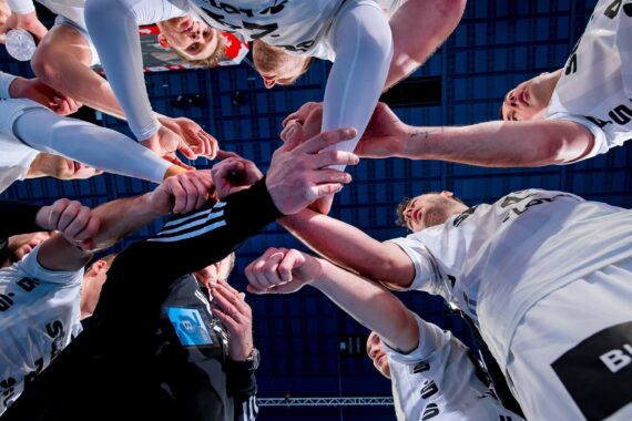 Eine Gruppe von Sportlern in weißen Sportuniformen drängt sich zusammen und hält die Hände in der Mitte zusammen, von unten gesehen vor einer blauen Decke, um Teamwork und Einigkeit vor einem Spiel zu zeigen.