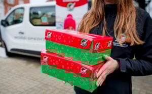 Eine Person hält zwei rote und grüne Schuhkartons mit weihnachtlichen Motiven und steht draußen neben einem weißen Lieferwagen. Die festlich geschmückten Kartons sind mit Logos versehen, und die Person trägt eine dunkle Jacke - bereit, Weihnachten zu feiern wie ein echter Handballstar.