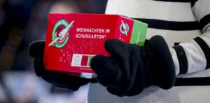 Eine Person in schwarz-weiß gestreifter Kleidung, die an einen THW Kiel Handball-Fan erinnert, hält einen rot-grünen Schuhkarton mit der Aufschrift "Weihnachten im Schuhkarton", der mit einem Logo und Sternen verziert ist - fertig als Kindergeschenk.