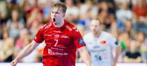 Ein männlicher Handballspieler in einem roten Trikot feiert leidenschaftlich auf dem Spielfeld, mit offenem Mund in einem Schrei, mit einem anderen Spieler in einem weißen Trikot verschwommen im Hintergrund. Die Szene ist energiegeladen und spielt während eines Handballspiels.