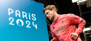 Ein männlicher Athlet in einer rosa Deutschland-Jacke steht neben einem blauen Schild mit dem Logo der Olympischen Sommerspiele Paris 2024.