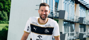 Ein Mann, der ein weiß-goldenes Sporttrikot mit Logos trägt, steht im Freien und lächelt in die Kamera. Hinter ihm befinden sich moderne Gebäude mit Balkonen und eine grüne, baumbestandene Fläche.
