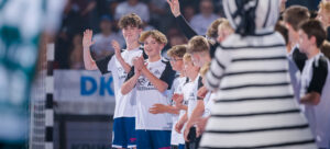 Eine Gruppe von Jungen in weißen Sporttrikots, die als THW Kiel Einlaufkinder agieren, stehen in einer Reihe und lächeln und winken. Im Vordergrund ist eine schwarz-weiß gestreifte Figur zu sehen, die das Nordderby gegen Flensburg zu sehen scheint.