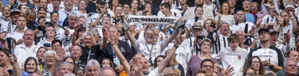 Eine große Menge begeisterter Fans, viele in weißen Hemden, jubeln und schwenken Schals bei einem Handballspiel des THW Kiel. Auf einem Schal steht WIR SIND KIEL und fängt die energiegeladene Atmosphäre der DHB-Pokal Saison 2024/2025 2. Runde.