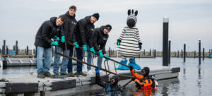 Eine Gruppe von sechs Personen in passenden Jacken und Handschuhen hilft zwei Tauchern, im Rahmen des THW Kiel För de Küste Cleanup Projekts Müll aus einem Yachthafen zu entfernen. Eine Person im Zebrakostüm steht hinter ihnen auf dem Steg. Im Hintergrund sind leere Stege und ruhiges Wasser zu sehen.