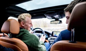 Zwei Männer sitzen auf den Vordersitzen eines Autos mit hellbraunem Lederinterieur und unterhalten sich über die kommende DHB-Pokal Saison 2024/2025. Der Fahrer lächelt seinen Beifahrer an, während er fährt, und auf dem Armaturenbrett ist ein GPS-Display zu sehen.
