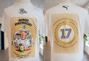 Ein weißes T-Shirt zu Ehren des Handballspielers Patrick Wiencek, das auf der Vorderseite sein Bild mit Mannschaftskameraden und "Ahoi Patrick!" zeigt. Auf der Rückseite ist die Nummer 17 zu sehen, umgeben von Jahreszahlen und Erfolgen in kreisförmigen goldenen Linien.