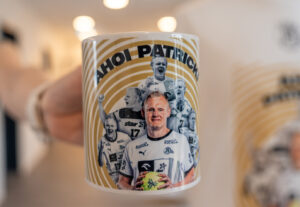 Nahaufnahme einer Tasse mit mehreren Bildern eines Mannes in einem weißen Sporttrikot, der einen grünen Ball hält, mit dem Text "AHOI PATRICK" über den Bildern und einem goldenen Kreismuster im Hintergrund.