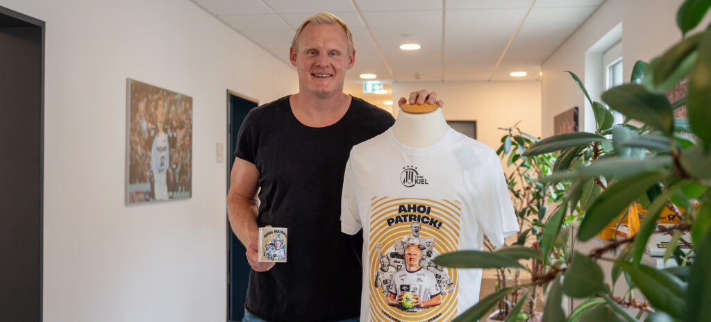 250714_Titel_Merch_Abschiedsspiel Ein lächelnder Mann steht drinnen neben einer Schaufensterpuppe, die ein weißes grafisches T-Shirt zeigt und eine passende Tasse hält. Das T-Shirt zeigt ein illustriertes Design mit Text und einem Porträt einer Person. Im Hintergrund sind Pflanzen und ein Hausflur zu sehen.