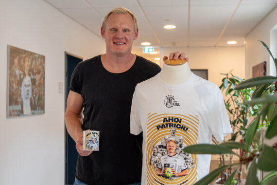 Ein lächelnder Mann steht drinnen neben einer Schaufensterpuppe, die ein weißes grafisches T-Shirt zeigt und eine passende Tasse hält. Das T-Shirt zeigt ein illustriertes Design mit Text und einem Porträt einer Person. Im Hintergrund sind Pflanzen und ein Hausflur zu sehen.
