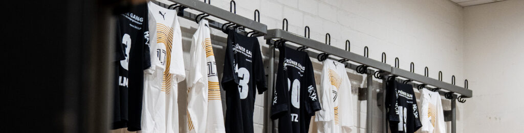 250725_Header_Versteigerung_2 Mehrere schwarz-weiße Fußballtrikots mit Nummern und Logos hängen ordentlich an Haken entlang einer Wand in einer Umkleidekabine, bereit für die Spieler, sie zu benutzen.