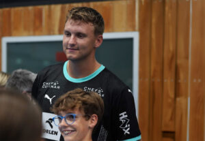 Ein lächelnder junger Mann in einem schwarzen Sporttrikot steht drinnen neben einem Jungen mit Brille und Zahnspange, im Hintergrund sind Holzplatten und ein Fenster zu sehen.
