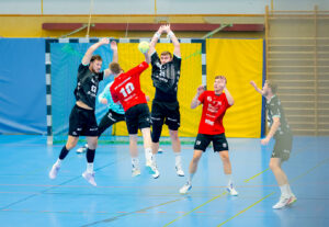 Zwei Handballspieler in schwarzen Trikots springen, um einen Schuss eines Spielers in einem roten Trikot zu blocken, der in der Luft ist und auf das Tor zielt, während zwei andere Spieler in einer Halle zusehen.