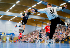 Ein Handballspieler in einem schwarzen Trikot springt und schießt auf das Tor, während ein Torwart in Weiß versucht, das Tor zu verhindern. Die Zuschauer füllen die Tribüne. Andere Spieler in schwarzen und roten Trikots sind auf dem Spielfeld zu sehen.