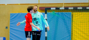 Zwei männliche Handballspieler, einer in einem roten Trikot und einer in einem blau-weißen Torwarttrikot, lächeln und umarmen sich auf einem Hallenplatz in der Nähe von Torpfosten und Netz.