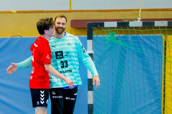 Zwei männliche Handballspieler, einer in einem roten Trikot und einer in einem blau-weißen Torwarttrikot, lächeln und umarmen sich auf einem Hallenplatz in der Nähe von Torpfosten und Netz.