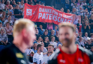 Eine Menschenmenge bei einer Sportveranstaltung hält ein großes rot-weißes Transparent mit der Aufschrift "DANKE FÜR 13 JAHRE PATENSCHAFT". Zwei Sportler stehen im Vordergrund, dicht dahinter sitzen die Fans.