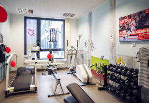 Ein kleiner, hell erleuchteter Fitnessraum mit Fitnessgeräten, darunter ein Laufband, ein Heimtrainer, Gewichte und ein Rudergerät. Sporttrikots und rote Luftballons schmücken den Raum, und ein Fenster lässt natürliches Licht herein.