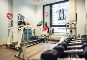 Ein moderner Fitnessraum mit einem Laufband, einem Rudergerät, Gewichten, roten Herzen, einem Sporttrikot an der Wand und großen Fenstern, die ein Stadtbild zeigen.