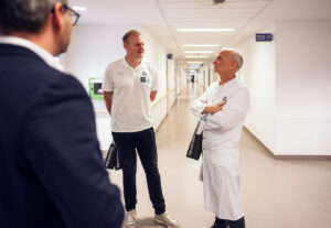 Drei Männer stehen in einem hell erleuchteten Krankenhausflur und unterhalten sich. Zwei von ihnen tragen weiße Hemden, einer ist als Arzt im Laborkittel gekleidet, und der dritte Mann trägt einen dunklen Anzug, den man von hinten sieht.