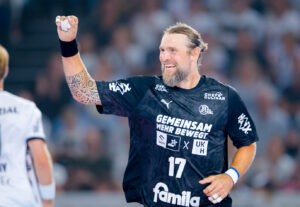 Ein männlicher Handballspieler in einem schwarzen Trikot mit der Nummer 17 und verschiedenen Sponsoren lächelt und hebt während eines Spiels jubelnd die Faust. Er hat einen Bart, lange zurückgebundene Haare und eine sichtbare Armtätowierung.