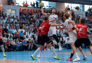 Ein Handballspieler in einem weißen Trikot springt, um den Ball zu werfen, während er von Spielern in roten und schwarzen Trikots in einer überfüllten Halle verteidigt wird. Das Publikum im Hintergrund schaut aufmerksam zu.