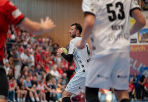 Ein Handballspieler in einem weißen Trikot hält den Ball und bereitet sich auf einen Spielzug vor, während im Hintergrund ein anderer Mitspieler und eine Menge Zuschauer in Rot zu sehen sind.
