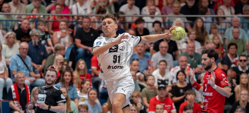 250729_Stralsund_THW_Titel Ein Handballspieler in einem weißen Trikot springt in die Luft, um den Ball zu werfen, während ein Gegner in einem roten Trikot zusieht. Im Hintergrund ist eine Zuschauermenge zu sehen, die das Spiel aufmerksam verfolgt.