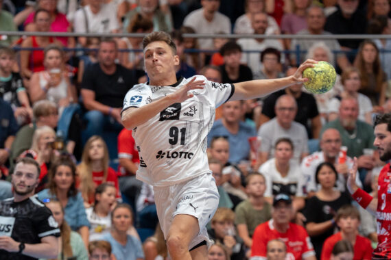 Ein Handballspieler in einem weißen Trikot springt in die Luft, um den Ball zu werfen, während ein Gegner in einem roten Trikot zusieht. Im Hintergrund ist eine Zuschauermenge zu sehen, die das Spiel aufmerksam verfolgt.