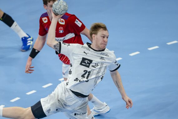 Ein Handballspieler in einem weißen Trikot springt in der Luft und bereitet sich auf den Wurf vor, während ein Verteidiger in einem roten Trikot versucht, ihn auf einem blauen Hallenplatz zu blockieren.
