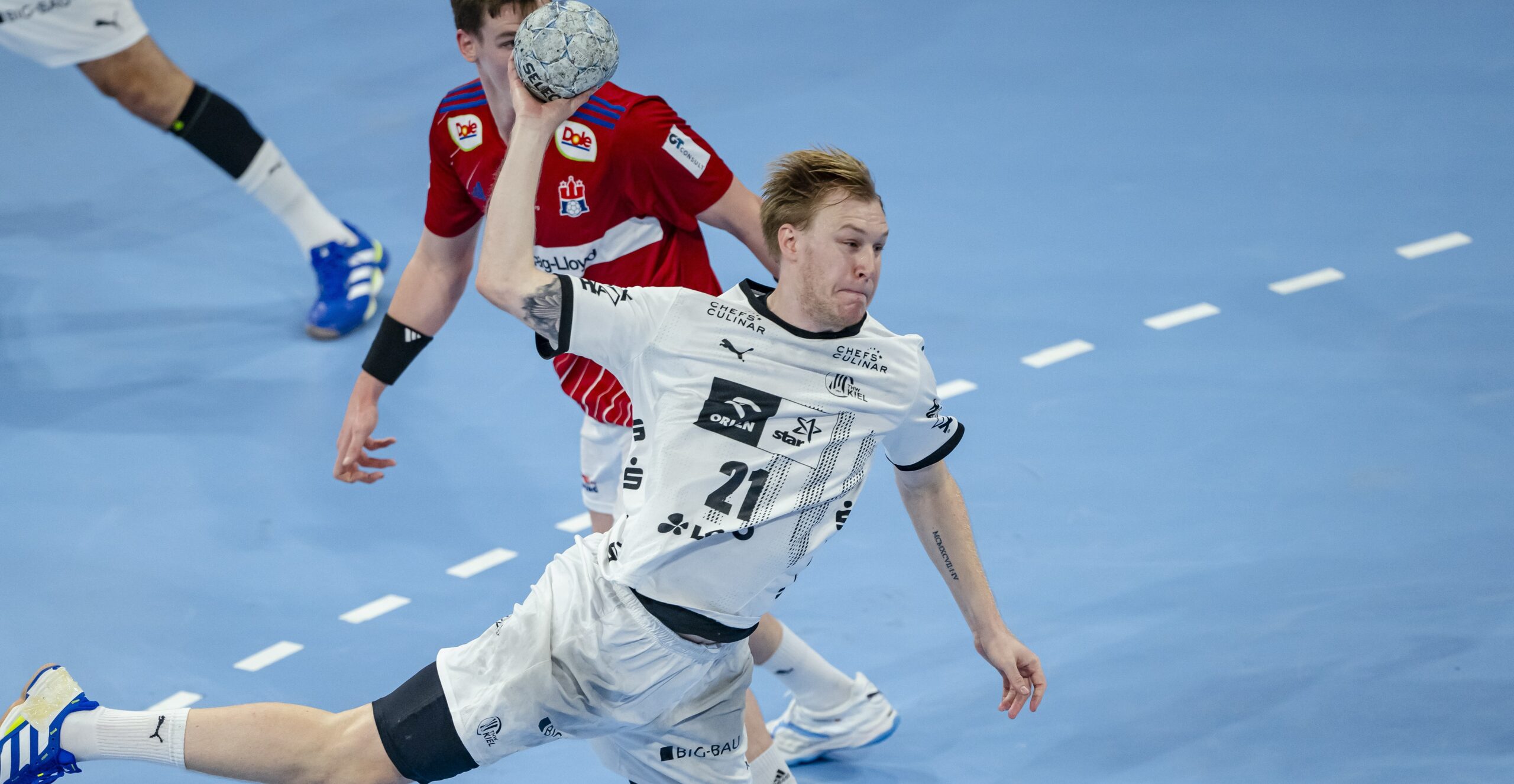 Ein Handballspieler in einem weißen Trikot springt in der Luft und bereitet sich auf den Wurf vor, während ein Verteidiger in einem roten Trikot versucht, ihn auf einem blauen Hallenplatz zu blockieren.