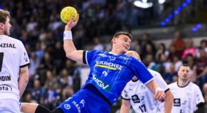 Ein männlicher Handballspieler in einem blauen Trikot bereitet sich darauf vor, einen gelben Ball zu werfen, umgeben von gegnerischen Spielern in weißen Trikots während eines intensiven Hallenspiels, mit einer jubelnden Menge im Hintergrund.