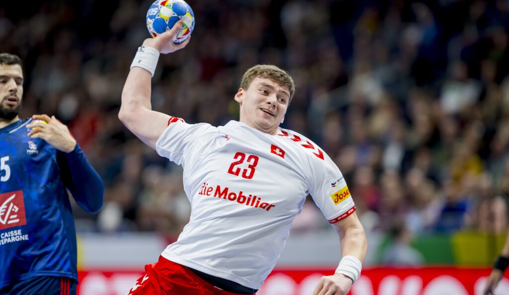 Ein männlicher Handballspieler in einem weißen Trikot mit der Nummer 23 bereitet sich darauf vor, den Ball während eines Spiels zu werfen, während im Hintergrund ein anderer Spieler in einem blauen Trikot zu sehen ist. Die Szene ist actionreich und spielt in einer Halle.