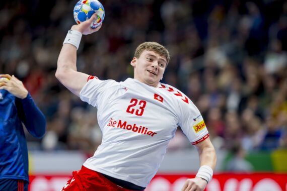 Ein männlicher Handballspieler in einem weißen Trikot mit der Nummer 23 bereitet sich darauf vor, den Ball während eines Spiels zu werfen, während im Hintergrund ein anderer Spieler in einem blauen Trikot zu sehen ist. Die Szene ist actionreich und spielt in einer Halle.