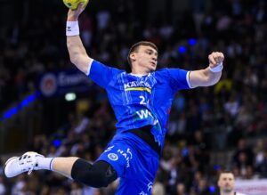 Ein männlicher Handballspieler in einem blauen Trikot, möglicherweise vom TVB Stuttgart, springt in der Luft und bereitet sich auf den Wurf vor, während der Handball-Bundesliga Saison 2025/2026, mit einer verschwommenen Zuschauermenge im Hintergrund.