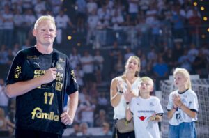 Ein blonder Sportler im schwarzen THW-Kiel-Trikot steht mit einem Mikrofon auf dem Sportplatz, hinter ihm klatschen eine Frau und zwei Kinder - der emotionale Abschied von Patrick Wiencek in der Saison 2025 in der hell erleuchteten Ostseehalle.
