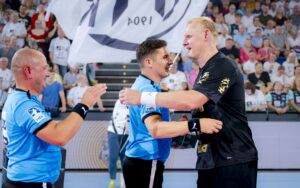 Ein Handballspieler in Schwarz lächelt und schüttelt einem Schiedsrichter in Blau auf dem Spielfeld während der THW Kiel Saison 2025 Abschied für Patrick Wiencek in der Ostseehalle die Hand, während ein anderer Schiedsrichter applaudiert und eine Menschenmenge mit einer großen Fahne den Hintergrund füllt.