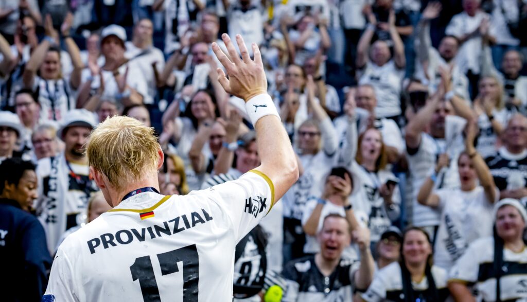 Eine blonde Sportlerin, die ein weißes Trikot mit der Nummer 17 trägt, winkt einer jubelnden Menge zu, die in passenden weißen und schwarzen Farben gekleidet in einem Stadion feiert.