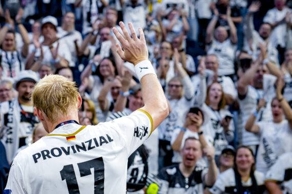 Eine blonde Sportlerin, die ein weißes Trikot mit der Nummer 17 trägt, winkt einer jubelnden Menge zu, die in passenden weißen und schwarzen Farben gekleidet in einem Stadion feiert.