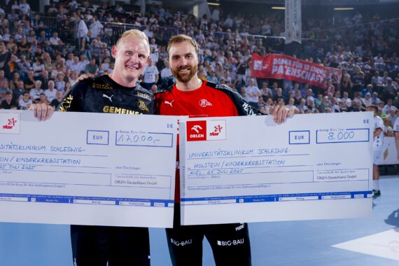 Zwei lächelnde Männer in Sportkleidung halten große Spendenschecks auf einem blauen Hallenplatz, mit einer jubelnden Menge im Hintergrund. Die Schecks sind auf 17.000 und 8.000 Euro für eine Kinderkrebshilfe ausgestellt.